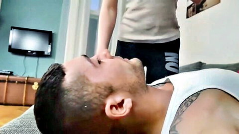Tattooed man in white tank top lies on couch as woman in beige top and black leggings stands over him teasingly