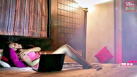 Seductive Indian woman in pink top lounging on bed with laptop, smiling erotically