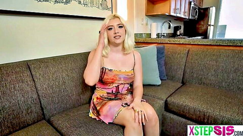 Young blonde stepsister teases with big tits cleavage in short colorful dress on couch