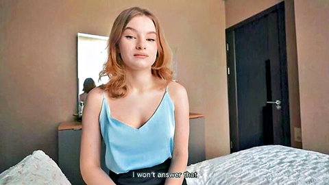 Skinny blonde babe in low-cut blue tank top sitting on bed looking reluctant