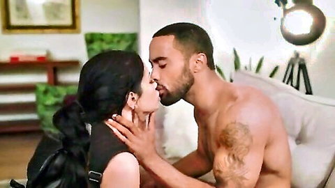 Busty brunette passionately kissing muscular tattooed black stud on couch