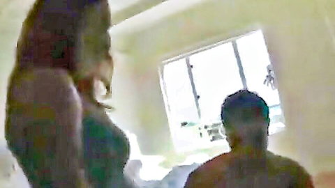 Busty Latina in tight green dress stands in hotel room over man on bed