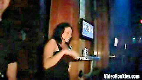 Busty brunette bartender in tight black dress flaunting deep cleavage while serving drinks at bar.