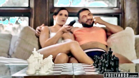 Brunette woman in tight white top spreads legs over chessboard on glass table while relaxing on couch with muscular man.