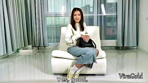 Cute Asian teen babe in sweater and skirt sitting on white chair smiling seductively