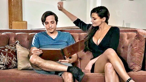Busty MILF in tight black dress flashes deep cleavage at young man reading leather album on couch