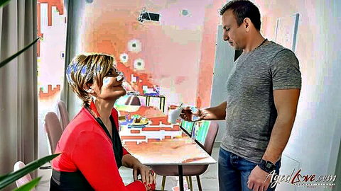 Mature blonde in red top smiles as man in gray shirt hands her coffee mug in art studio