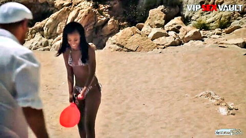 Hot ebony babe in skimpy white bikini holds red beach paddle smiling back at guy grabbing her on sunny rocky beach