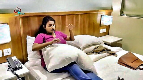 Busty South Asian woman in pink top and jeans gesturing on hotel bed
