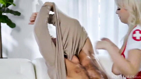 Blonde nurse in red uniform pulls muscular man's shirt off exposing his ripped torso.