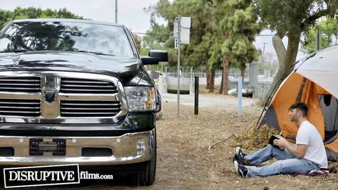 Bearded man in white shirt sits by orange tent holding camera next to black Ram truck outdoors