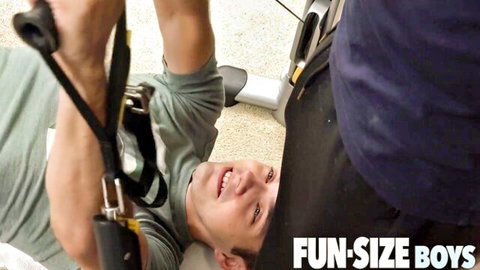 Smiling muscular twink in gray tank top looks up at daddy's crotch bulge in shorts on floor