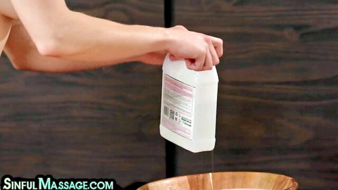 Hand pouring slippery massage oil from jug into wooden bowl in sensual massage room