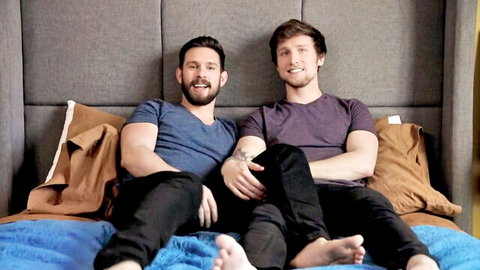 Two handsome young gay men sitting barefoot on bed smiling intimately