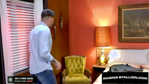 Handsome man in white shirt and jeans standing in retro orange bedroom on Raging Stallion.