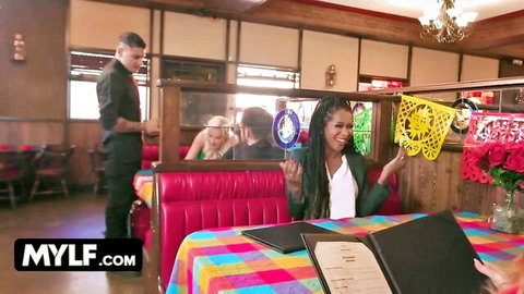 Smiling dreadlocked MILF holds yellow papel picado in colorful restaurant booth with blonde and waiter