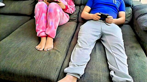 Stepsister in pink pajamas sits with stepbrother gaming, barefoot tease
