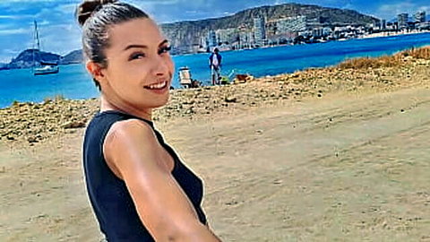 Brunette woman smiling in black tank top taking beach selfie with ocean and city view