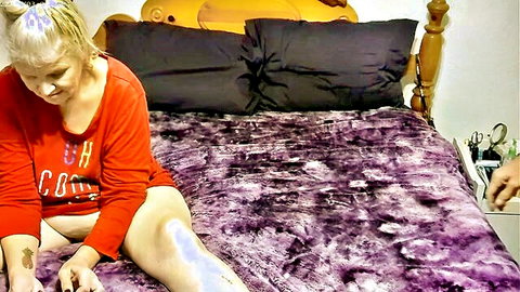 Mature blonde granny in red OH shirt sits spread-legged on purple bed