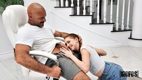 Muscular bald guy embraces petite redhead teen on his lap in crop top and shorts