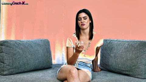 Curvy brunette in sheer top and tiny denim shorts sits teasing on couch with legs apart