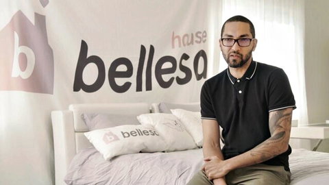 Tattooed man with glasses sits on bed in Belle House studio bedroom set.