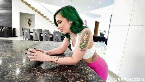 Gia Paige with green hair and tattoos leans seductively in white tank top and pink leggings POV