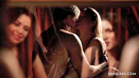 Muscular shirtless man passionately kissing blonde woman surrounded by smiling semi-nude girls in red curtains