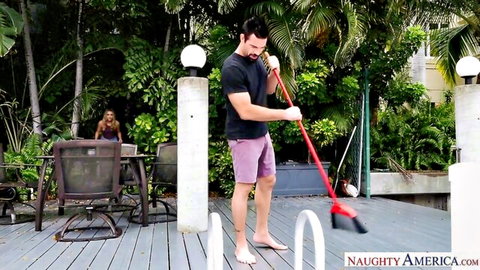 Muscular guy in shorts sweeping outdoor deck while sexy long-haired wife watches from plants