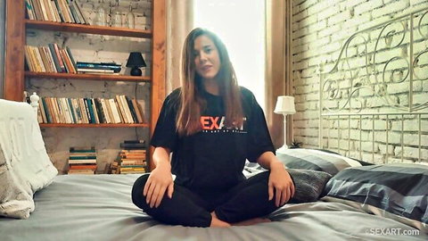 Sybil in casual black SEXART t-shirt, sitting cross-legged on bed for MetArt behind-the-scenes