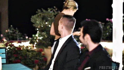 Blonde woman in brown dress stands between two suited men on dark patio night scene