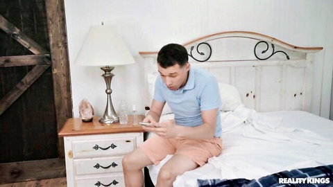 Clothed man in blue shirt and shorts sitting on bed checking phone in bedroom