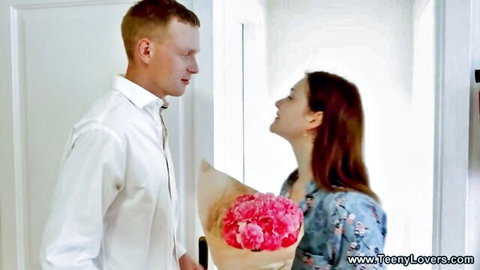 Handsome man in white shirt gives pink peonies bouquet to smiling petite babe in floral dress at doorway