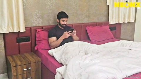 Muscular Desi husband in black shirt sits on bed holding phone, ready for wife action