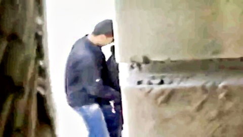 Blurry man in beanie and jacket bends over peeking behind gray stone wall in snowy outdoor setting