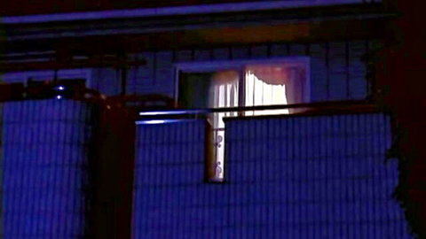 Lit window in house at night with sheer curtain partially drawn revealing warm interior glow