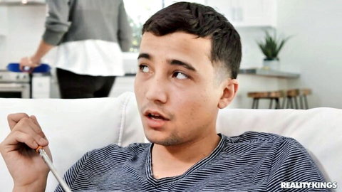 Surprised young guy smoking on couch while brunette MILF cooks in kitchen background