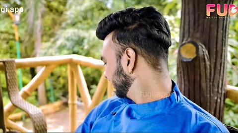 Handsome bearded Indian man in blue kurta sitting on outdoor wooden bench with green trees