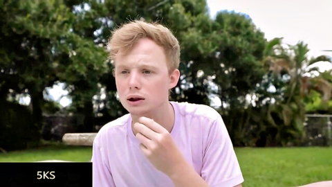 Freckled blonde twink in pink shirt gestures finger to open mouth outdoors in grassy yard