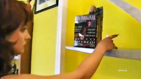 Brunette with short hair points at Rocco Siffredi original book on yellow shelf in vintage porn scene