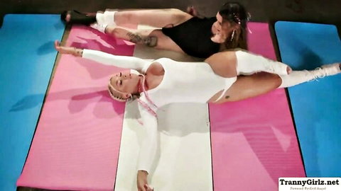 Blonde trans in tight white bodysuit stretches legs wide on yoga mats with brunette beside her