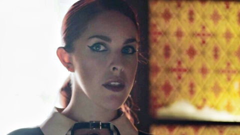 Sultry redhead with heavy eyeliner and choker collar smirks seductively in dramatic lighting