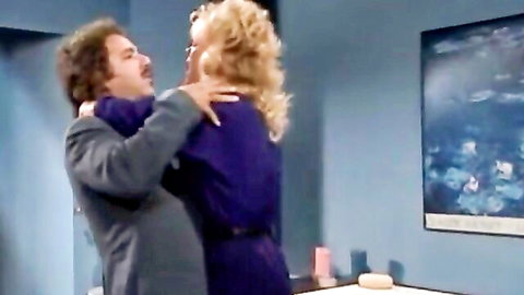 Blonde in purple dress embraced from behind by curly-haired man in suit