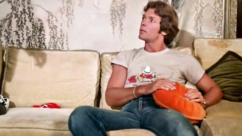 Vintage hunk in tight jeans and t-shirt lounges on couch holding orange pillow over crotch