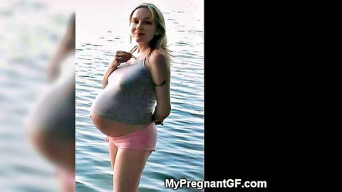 Blonde pregnant woman posing in gray tank top and pink shorts by water
