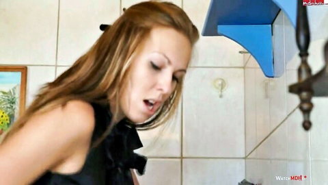 Blonde woman with small tits leans forward with open mouth in kitchen POV tease