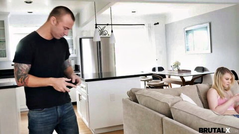 Tattooed guy texts in kitchen while petite blonde babe lounges on couch in tight pink top