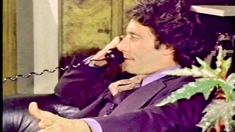 Curly-haired man in purple suit talking on retro phone gesturing thumbs up on leather couch