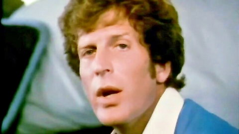 Close-up of curly-haired 70s man in blue shirt singing with mouth open