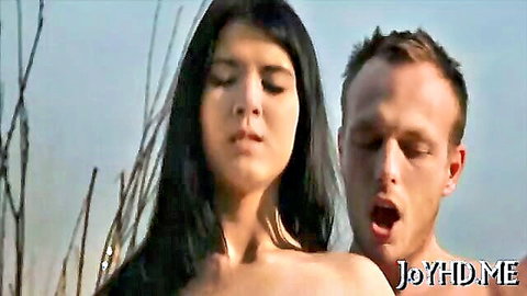 Brunette teen topless outdoors with man behind, intense expression